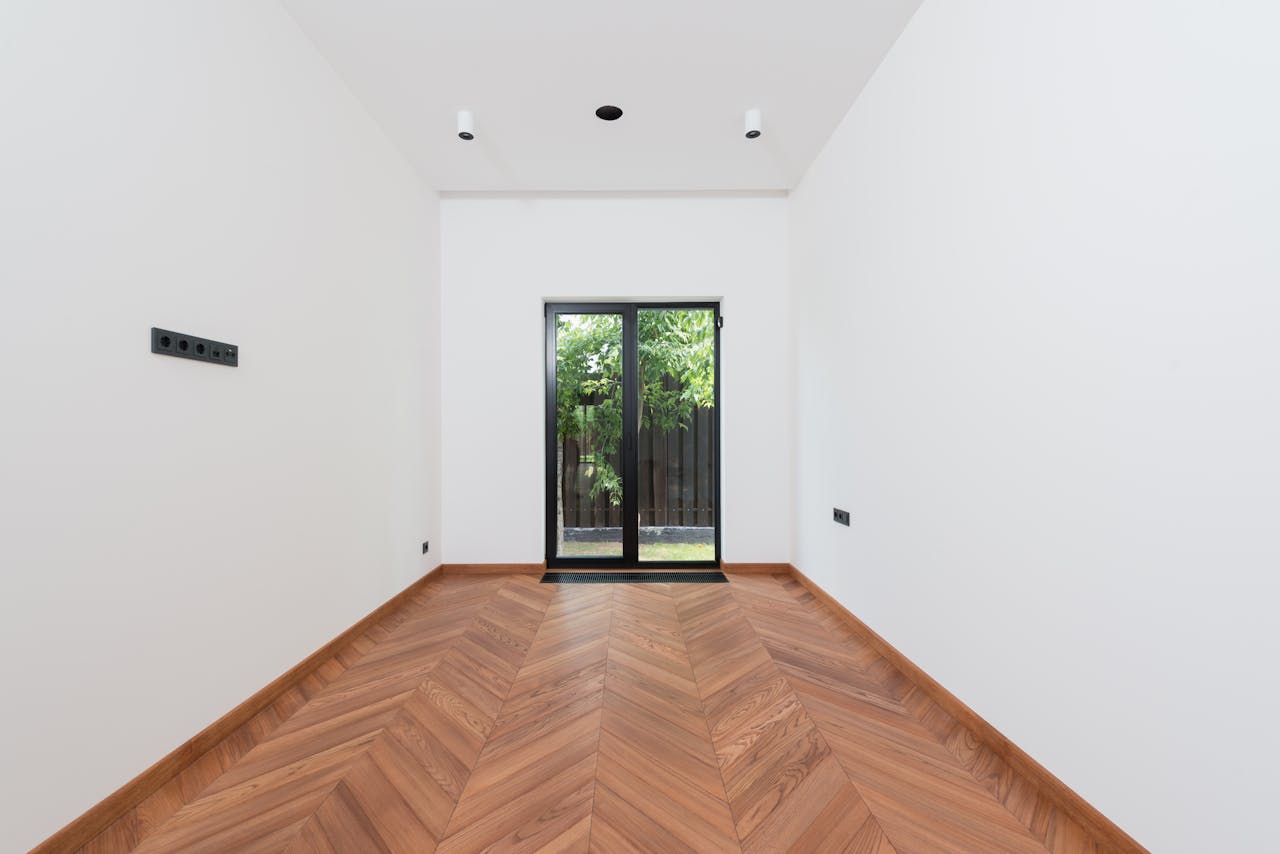 about-img A clean, minimalist interior room with white walls, wooden parquet flooring, and a glass door.